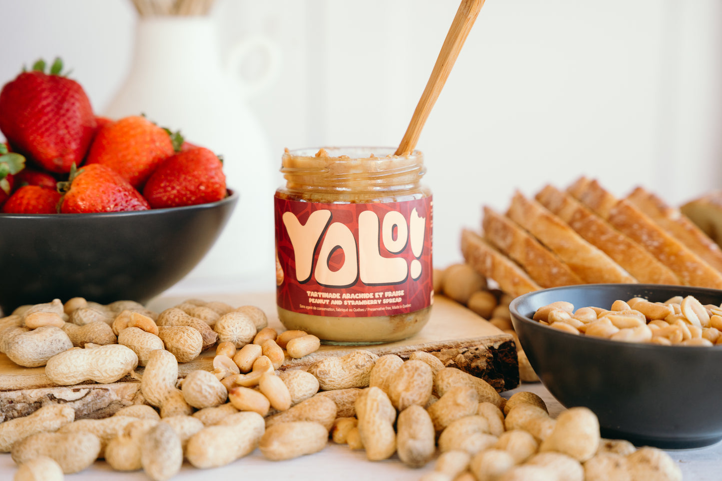 Tartinade arachide et fraise avec un couteau qui trempe dedans le pot. Un fond aveec des arachides, cacahouètes, fraises dans un bol et tranches de pain