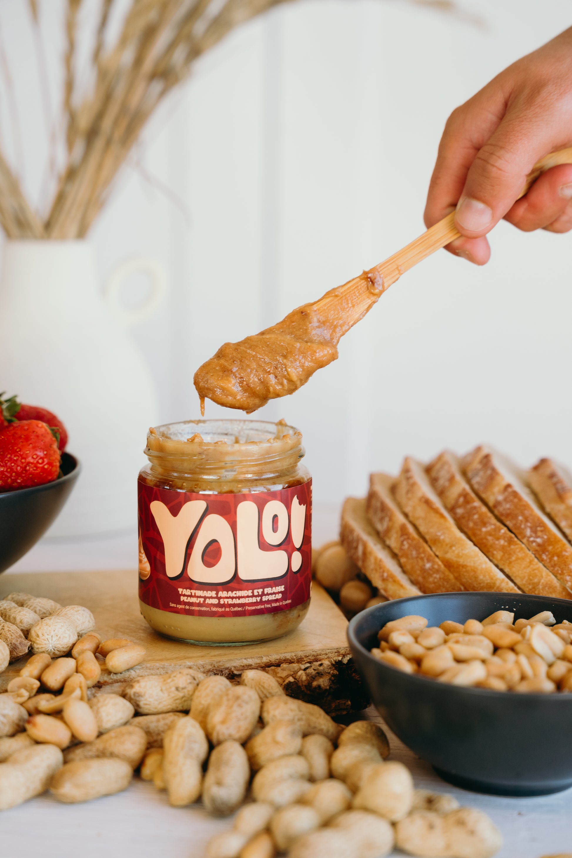 Tartinade arachide et fraise avec un couteau qui trempe dedans le pot. Un fond aveec des arachides, cacahouètes, fraises dans un bol et tranches de pain