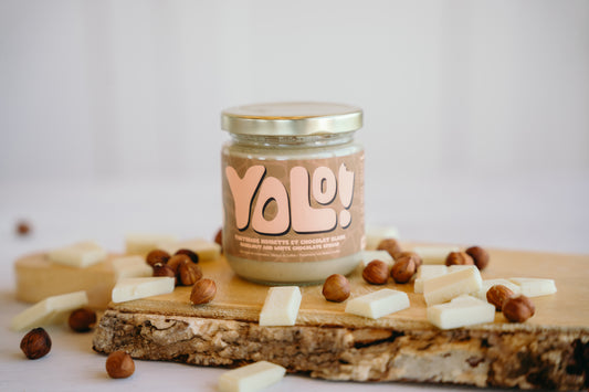 Pot de Tartinade de noisette et chocolat blanc 3 de Yolonuts sur une planche de bois rustique et des noisettes et morceaux de chocolat blanc