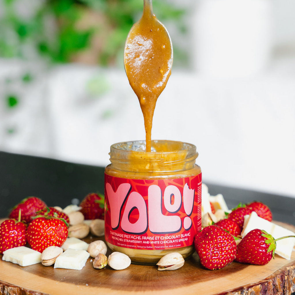 Pot de Tartinade de pistaches et chocot blanc et fraise avec une cuillère qui récolte son contennu entouré de fraise, chocolats blanc en morceaux et des pistaches en écaille sur une planche de bois