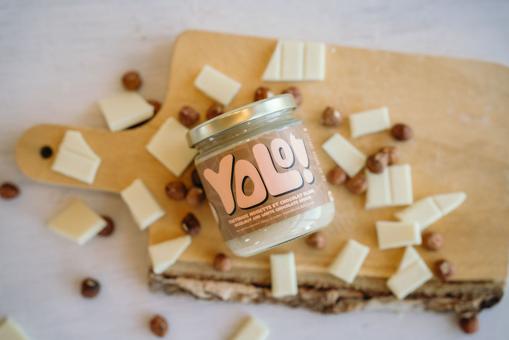 un pot de Tartinade noisette et choco blanc couché sur une planche à découpée avec des morceaux de chocolat blanc et des noisettes