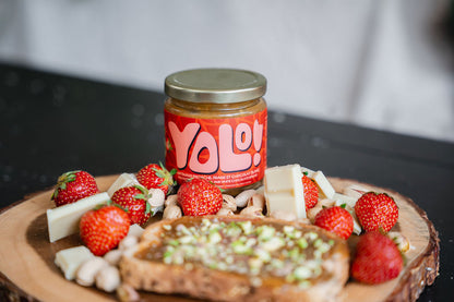 un pot de tartinade fraise pistache chocolat blanc sur une planche de tronc d'arbre entourné de fraises, morceaux de chocolat blanc, des pistaches et une roties avec de la tartinade et des pistaches en morceaux
