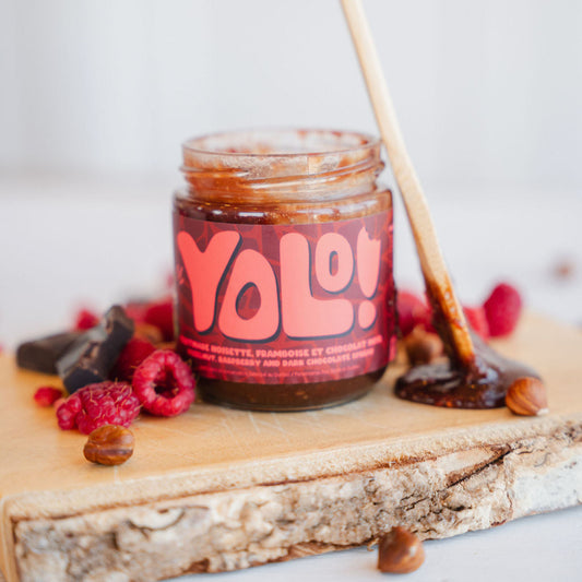 Pot de tartinadeframboise noisette chocolat noir sur une planche de bois avec des framboise et du chocolat noir à côté avec une cuillère couverte de tartinade acoté sur le pot
